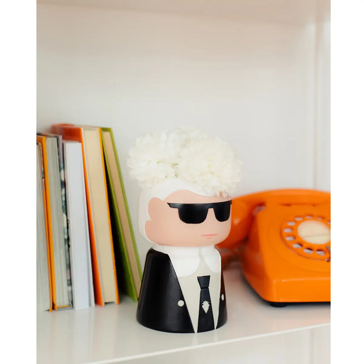 Vaso a forma di Karl Lagherfeld, con fiori bianchi. Sullo sfondo libreria bianca, libri e telefono
