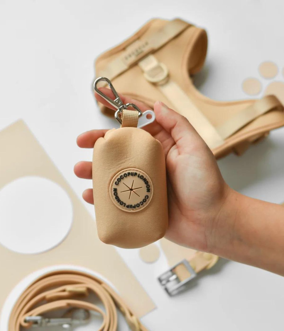 Porta Sacchetti colore biscotto tenuto in mano, con collare e guinzaglio abbinati sullo sfondo.