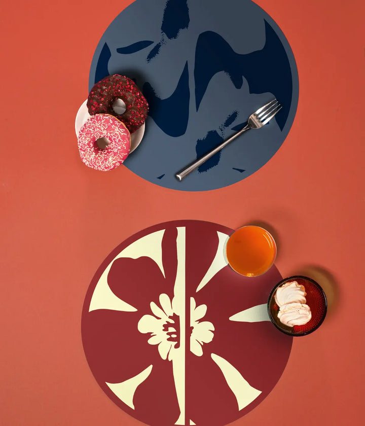 Tavolo arancione con due tovagliette tonde: blu e rosso e verde con fiore. Ciambelle e forchetta.