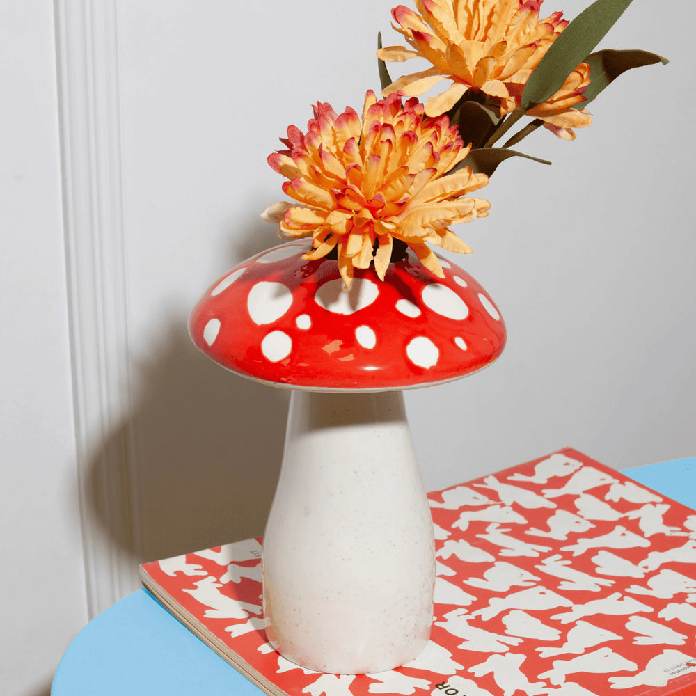 Vaso a forma di fungo amanita rosso con fiori gialli, appoggiato su un libro rosso e tavolo azzurro.