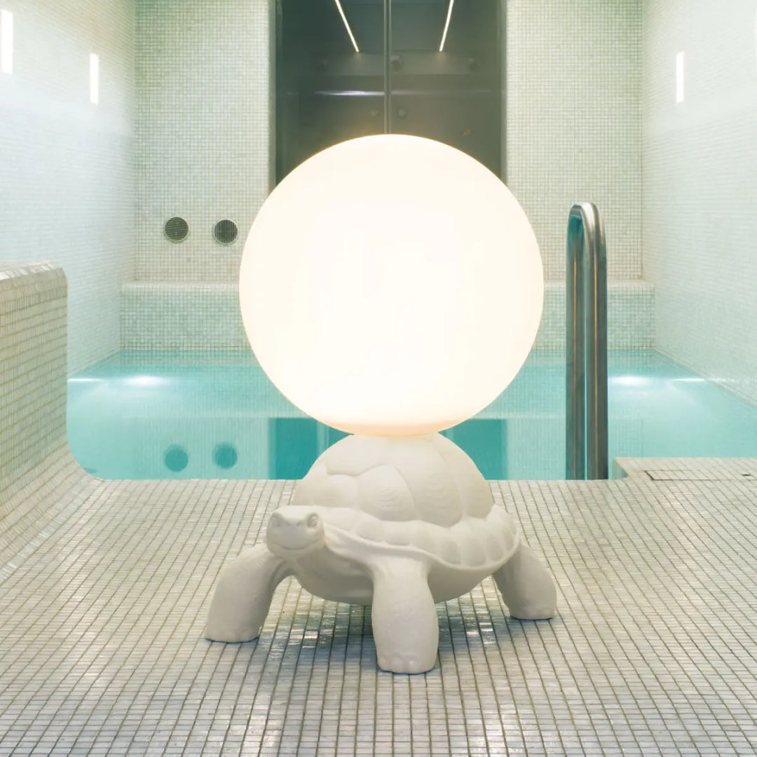Lampada da terra a forma di tartaruga bianca con sfera luminosa sul carapace Qeeboo posizionata sul bordo di una piscina interna.