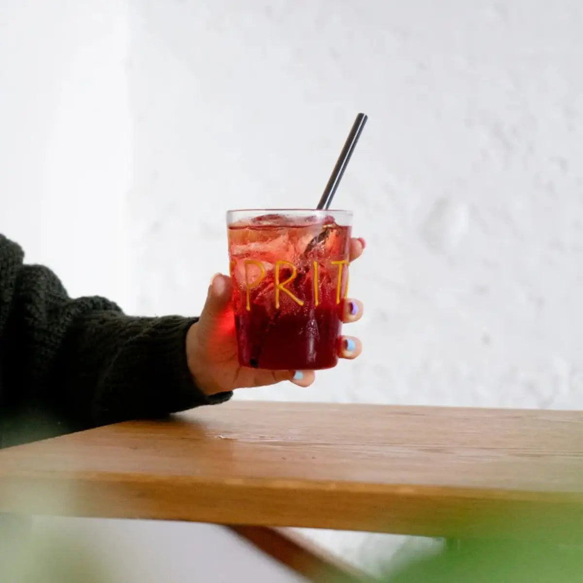 Mano che tiene il bicchiere di vetro trasparente con scritta gialla in rilievo "Spritz". tavolo di legno