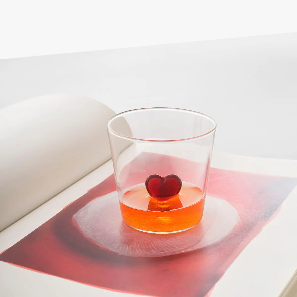 Bicchiere trasparente in vetro con cuore in vetro rosso sospeso al centro. Il bicchiere è appoggiato su un libro aperto