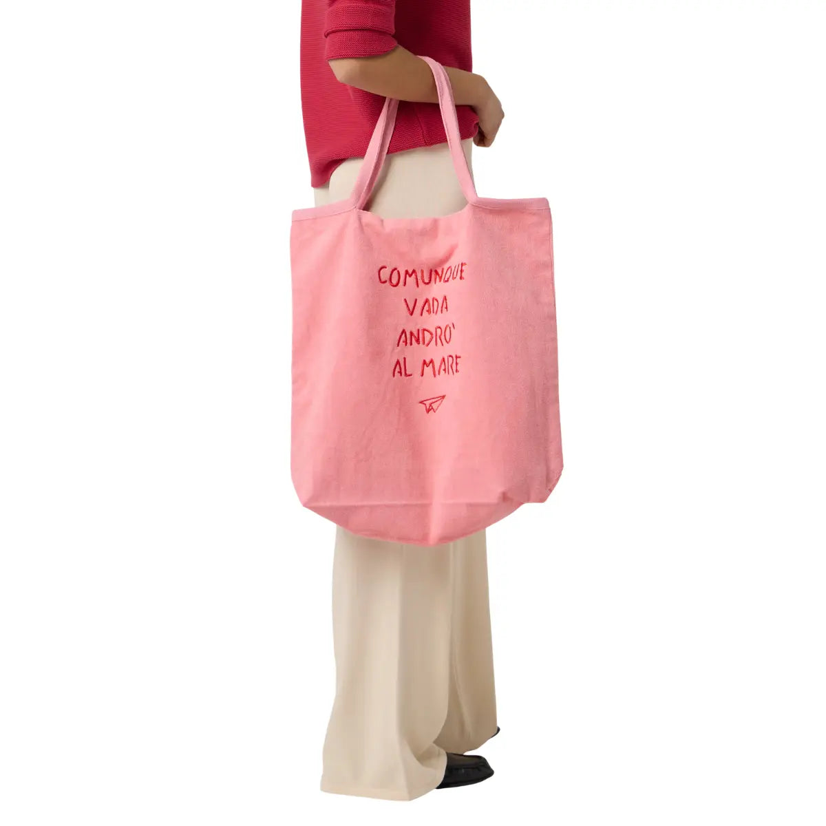 Donna che tiene appeso al braccio la borsa rosa da mare con scritta "comunque vada andrò al mare". Sfondo bianco 