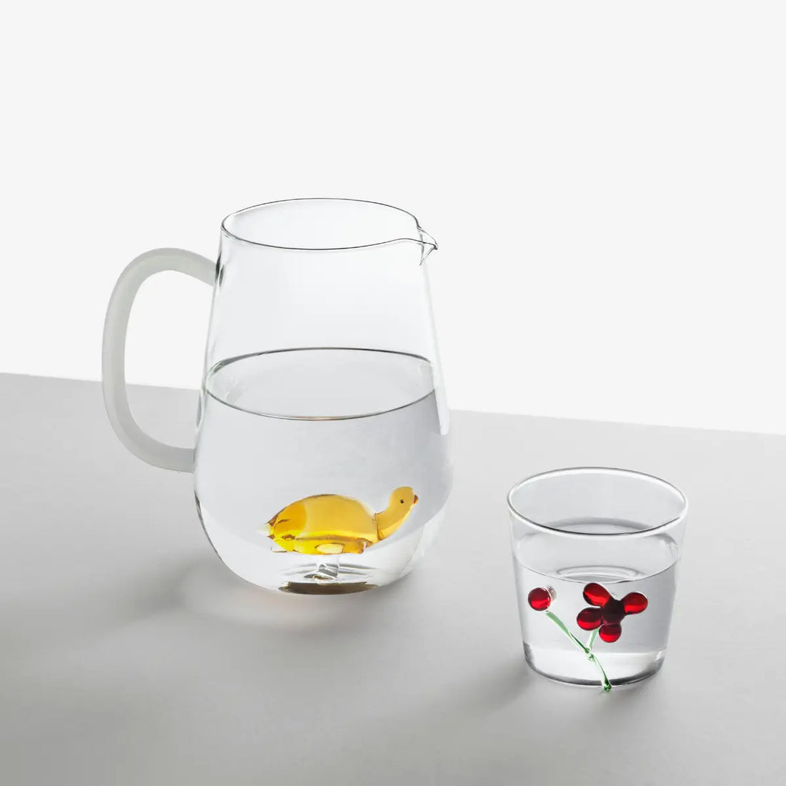 Brocca di vetro con una tartaruga di vetro giallo al centro e un bicchiere con decorazione rossa. entrambe sono appoggiate su un tavolo