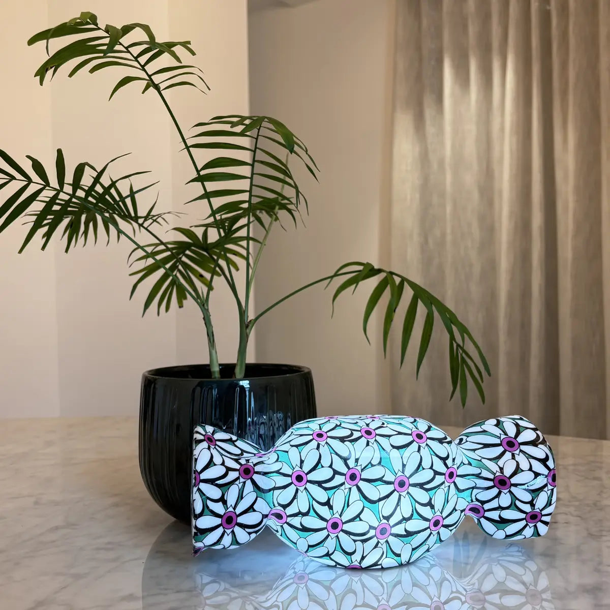 Lampada da tavolo a forma di caramella con base verde e fantasia con fiori bianchi con al centro degli occhi fucsia. appoggiata su un tavolo di marmo con dietro una pianta su vaso nero.