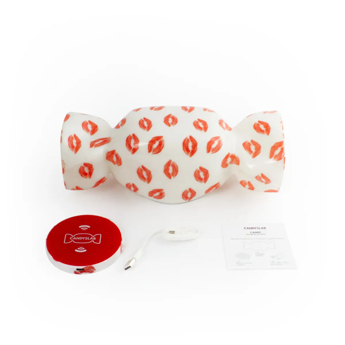 Contenuto confezione della lampada da tavolo a forma di caramella con base bianca e fantasia con baci rossi. Oltre la lampada c'è la base di ricarica wireless, un cavo di ricarica e il libretto delle istruzioni. Sfondo bianco 