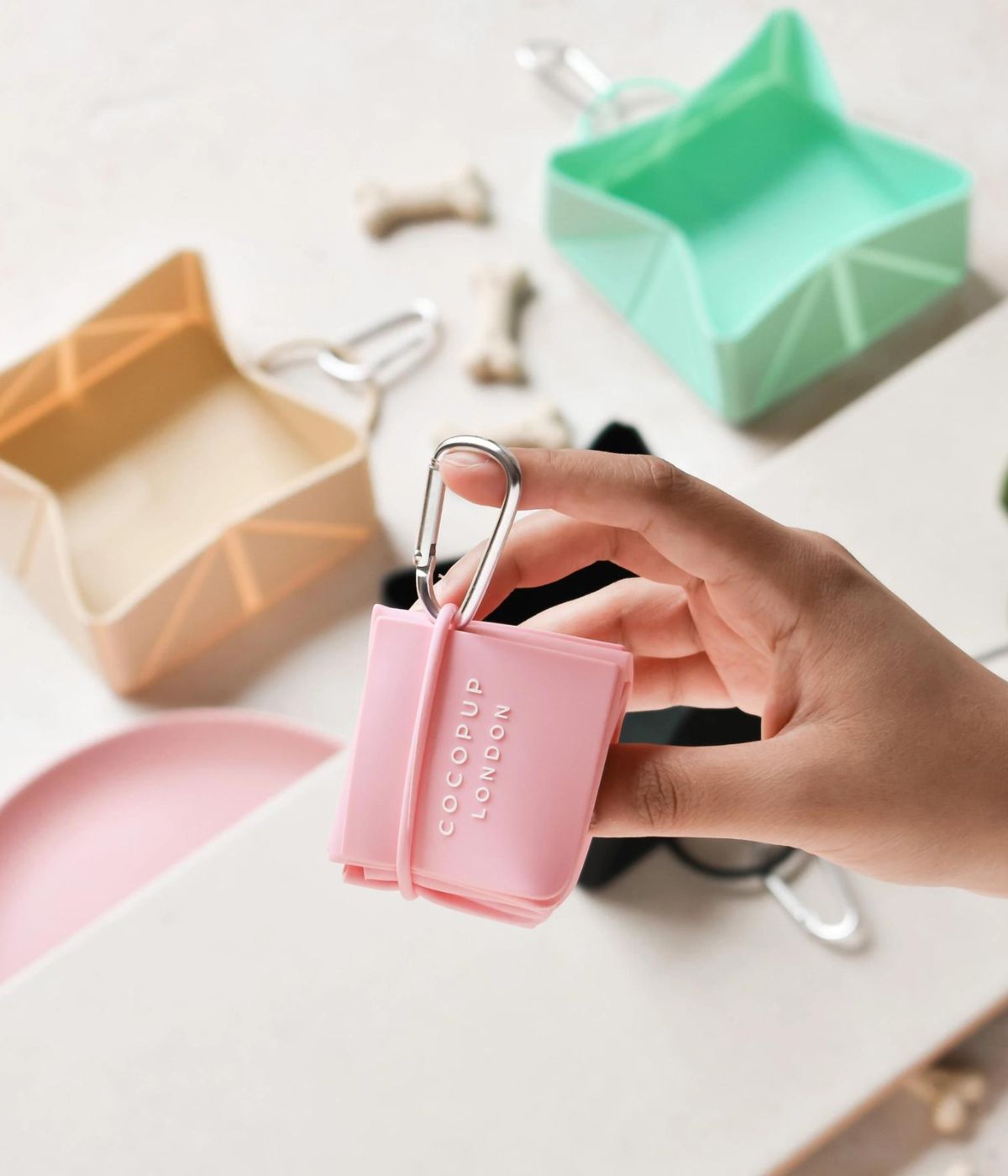 Mano che tiene la ciotola da viaggio pieghevole rosa con altre ciotole colorate sullo sfondo.