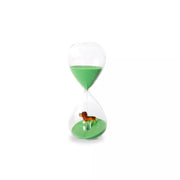 Clessidra in vetro con sabbia verde. All'interno della clessidra è presente un piccolo bassotto marrone in vetro. Sfondo bianco 