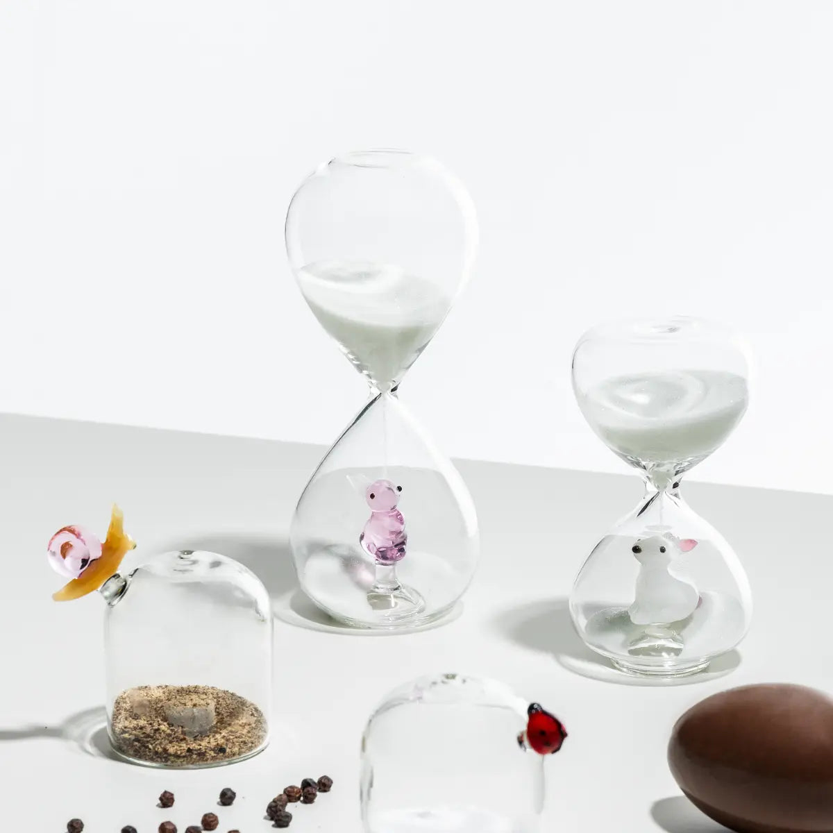 Clessidra con coniglio rosa sospeso, clessidra con coniglio bianco e altri elementi della collezione Animal Farm di Ichendorf appoggiati su un tavolo con sfondo bianco