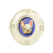 Cornice bianca di forma ovale con il volto di un bulldog in rilievo con gli occhi azzurri e a macchie bianche e marroni su sfondo viola. L'immagine è su sfondo bianco.