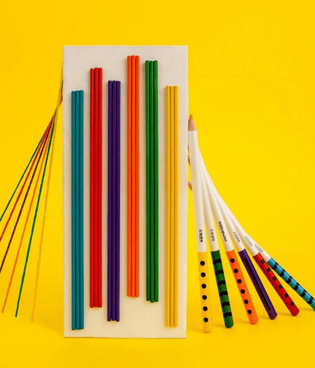 Rettangolo con righe colorate e matite appoggiate lateralmente. Immagine su sfondo giallo.