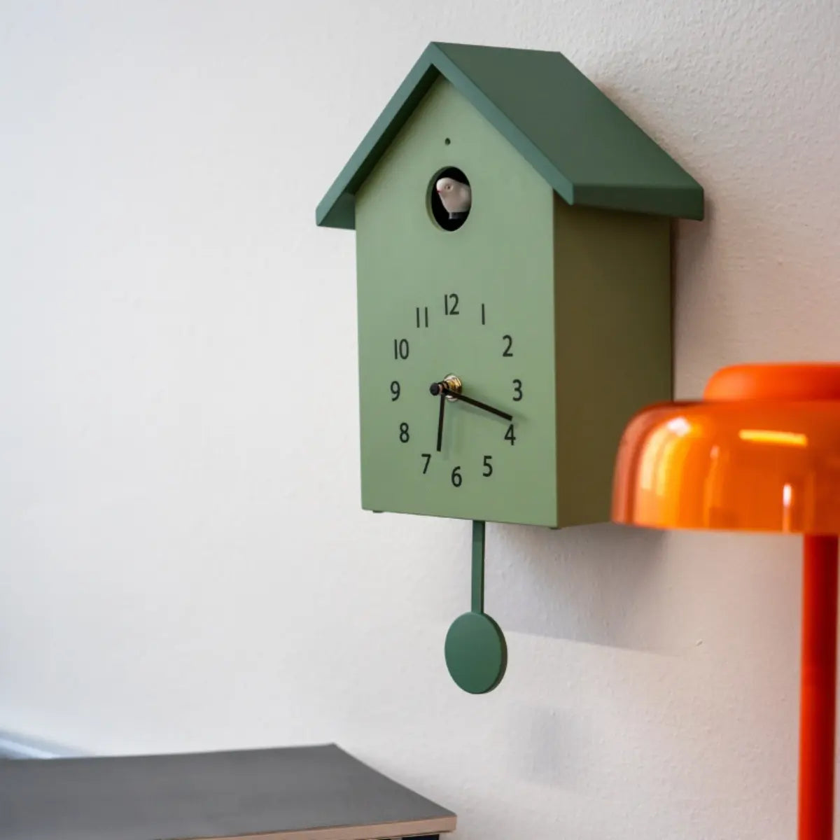 Cucu a forma di casetta verde appeso su un muro bianco. Si intravede una lampada arancione in primo piano 