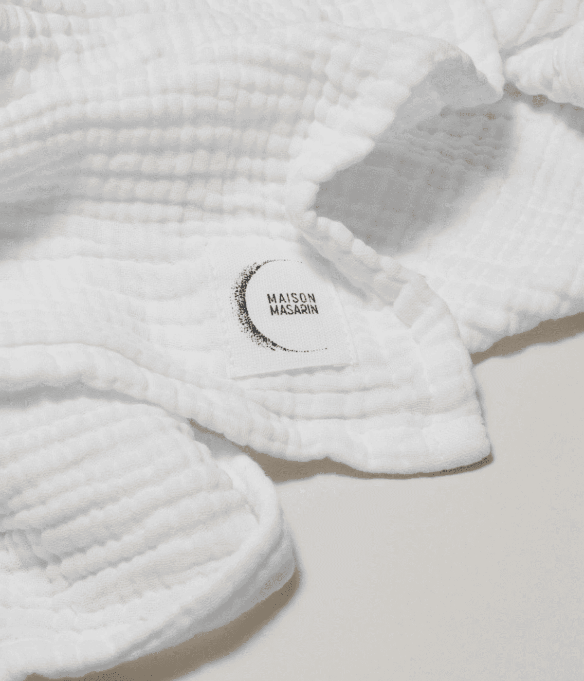 Inserto in tessuto con scritta Maison Masarin, cucito su asciugamano bianco in garza. Sfondo bianco.