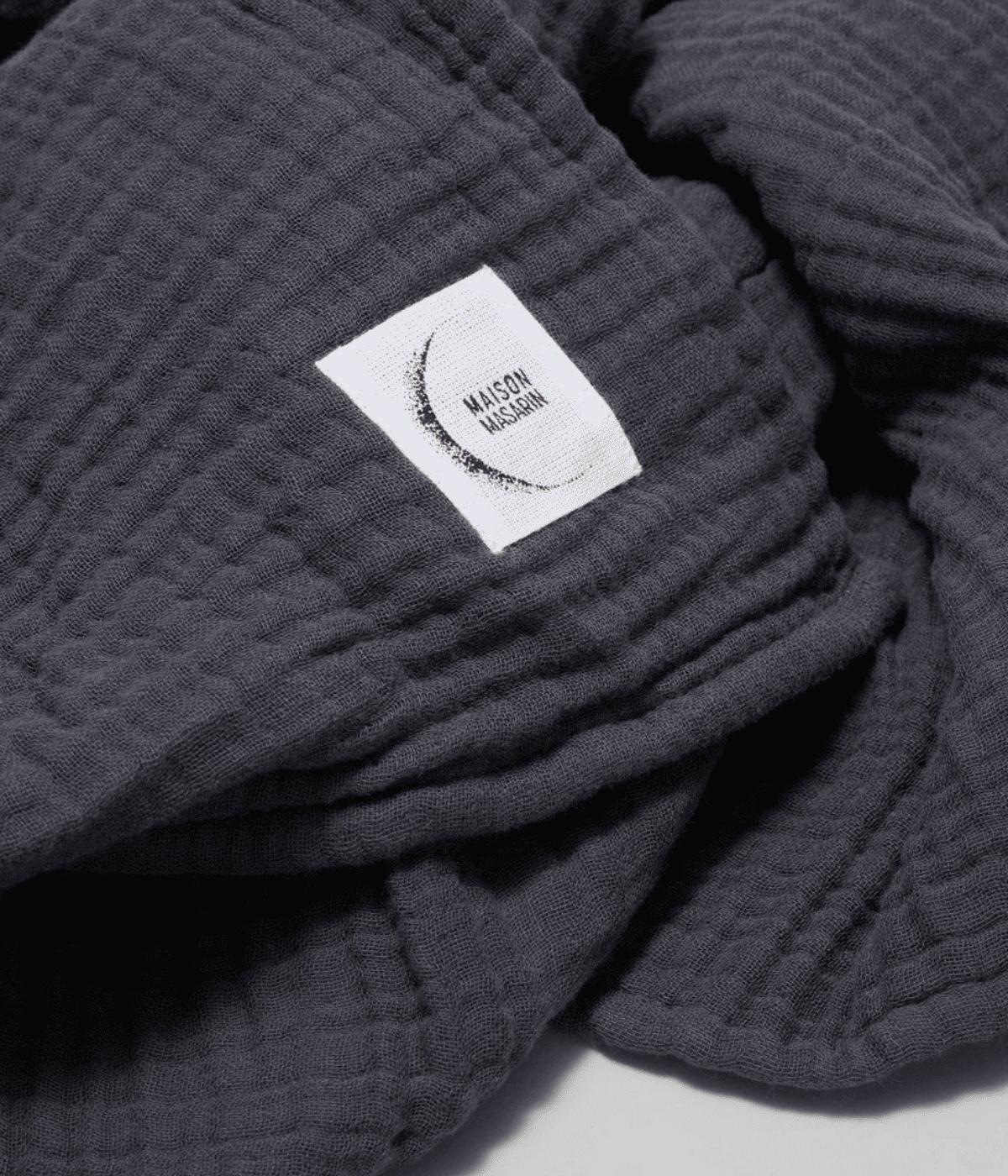 Inserto in tessuto con scritta Maison Masarin, cucito su asciugamano blu scuro in garza. Sfondo bianco.