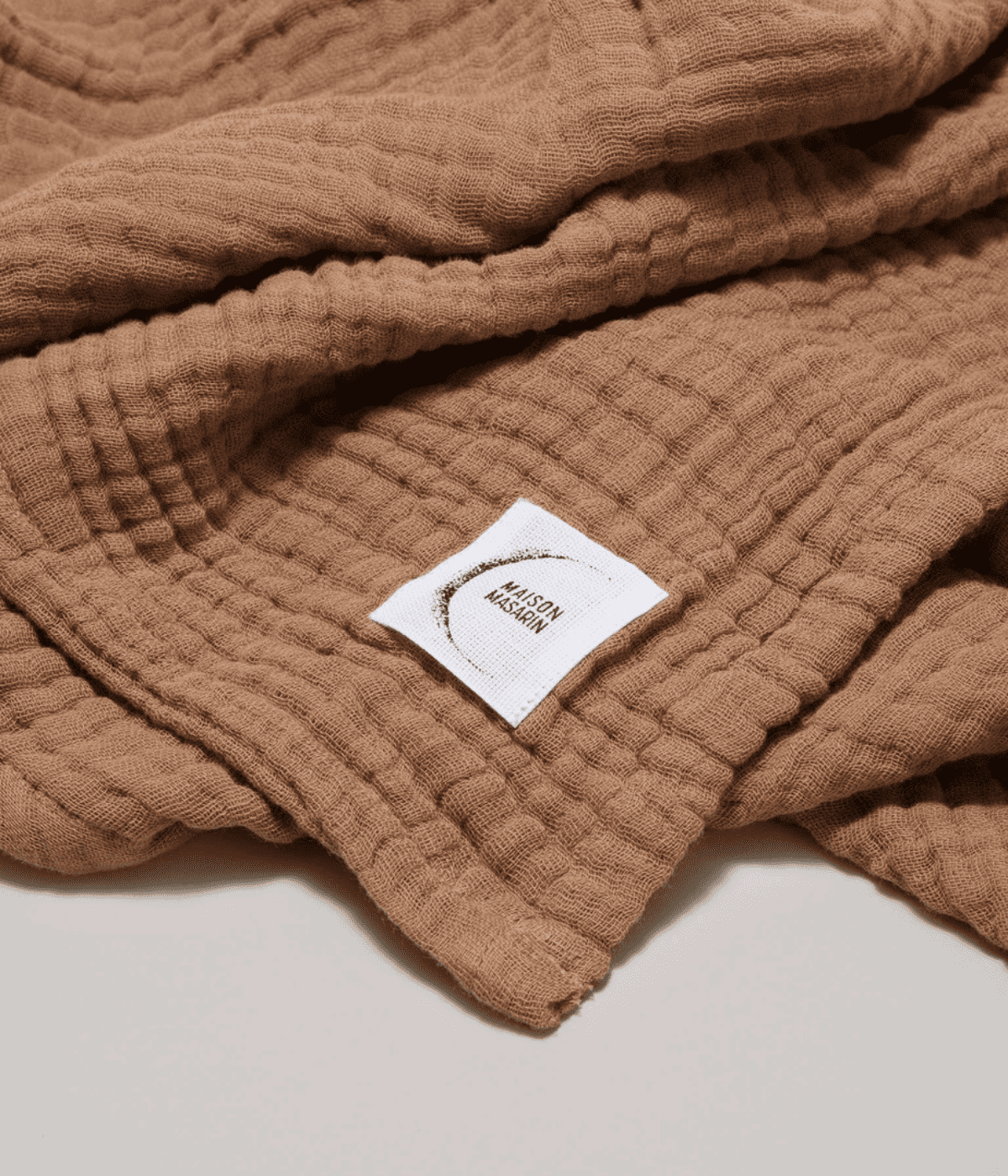 Inserto in tessuto con scritta Maison Masarin, cucito su asciugamano Tabacco in garza. Sfondo bianco.