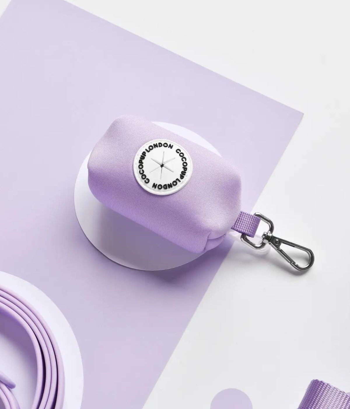 Porta sacchetti lilla con moschettone, marchio rotondo bianco con scritta Cocopup. Sfondo lilla