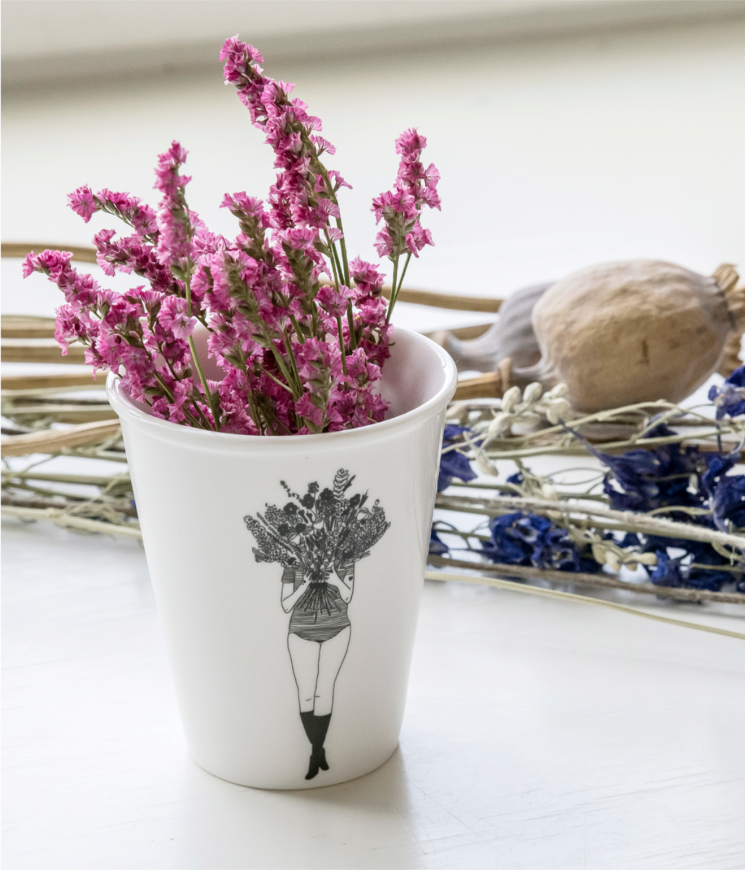 Fiori rosa dentro tazza bianca con disegno nero di donna con mazzo di fiori. Sfondo tavolo e fiori.