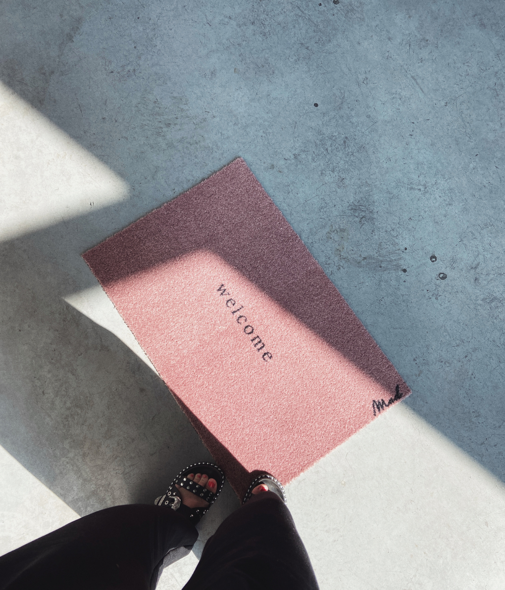 Persona che guarda dall'alto lo zerbino rosa con scritta nera Welcome. Sfondo pavimento