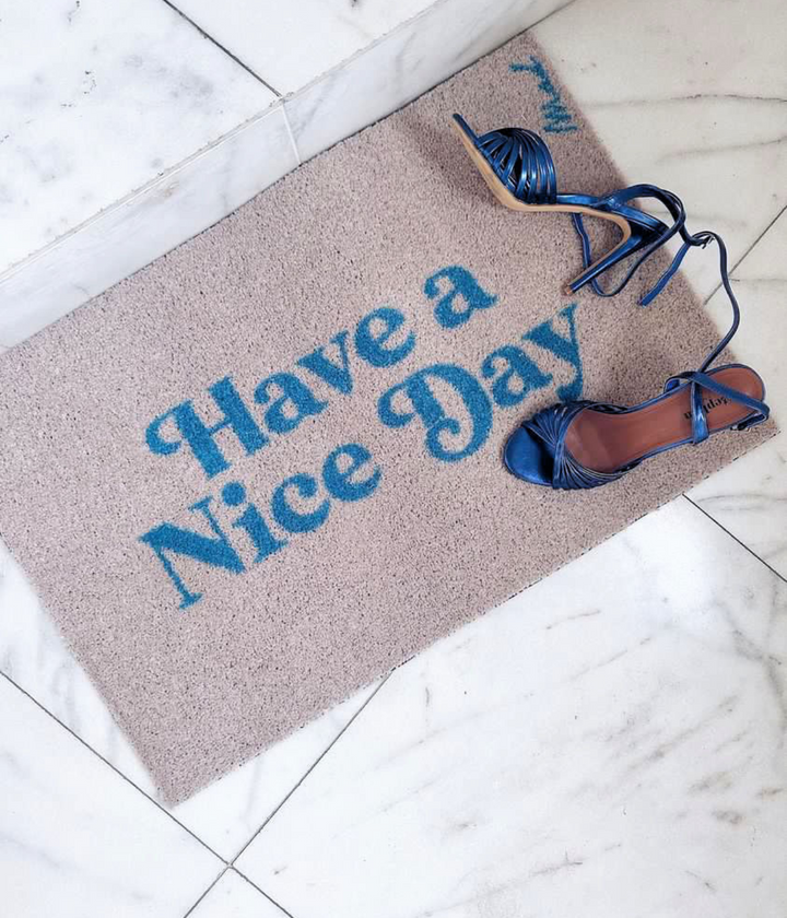 Zerbino beige con scritta azzurra Have a Nice Day. Scarpe appoggiate sopra. Sfondo pavimento marmo