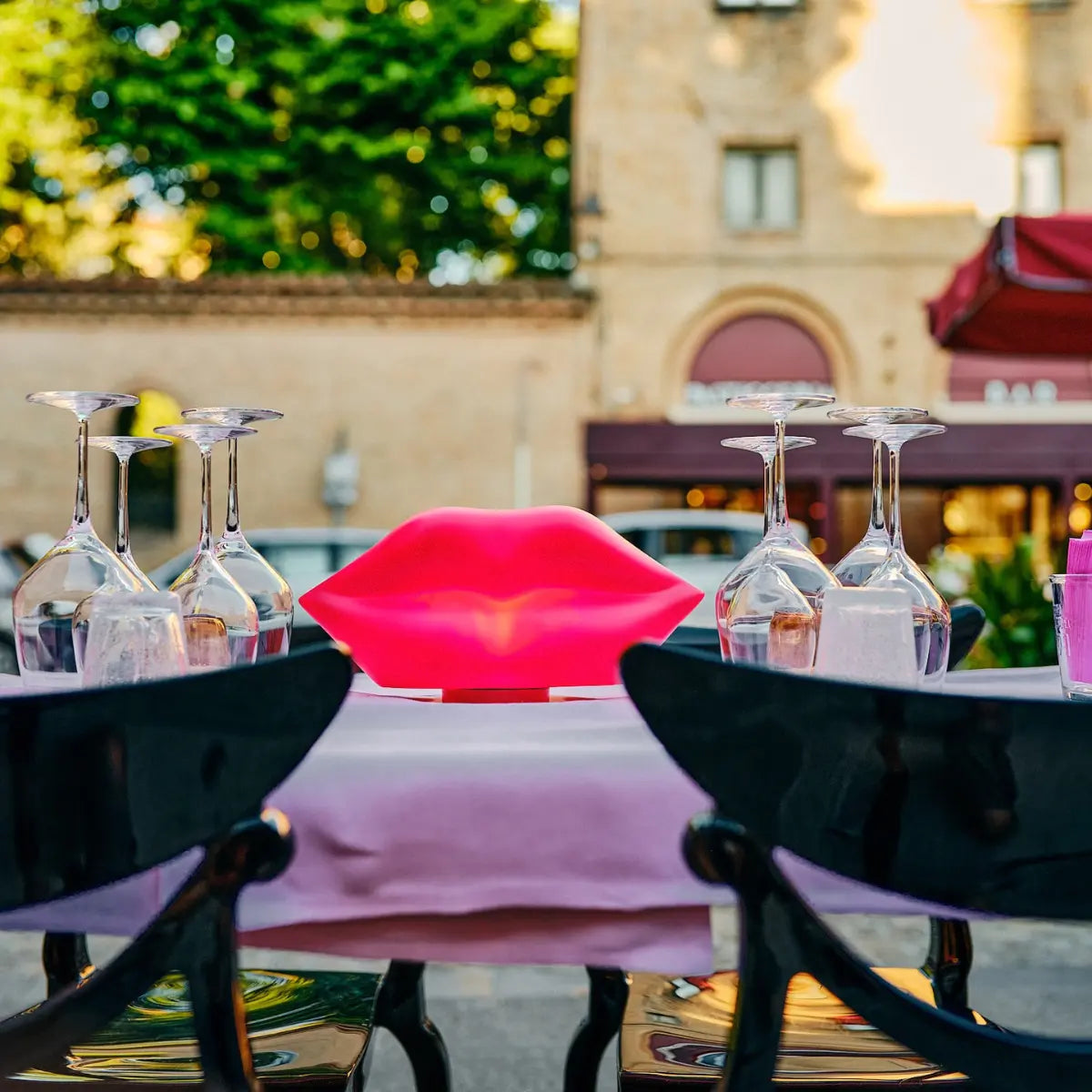 Lampada da tavolo fucsia a forma di labbra. Appoggiata su tavolo apparecchiato. Sfondo città