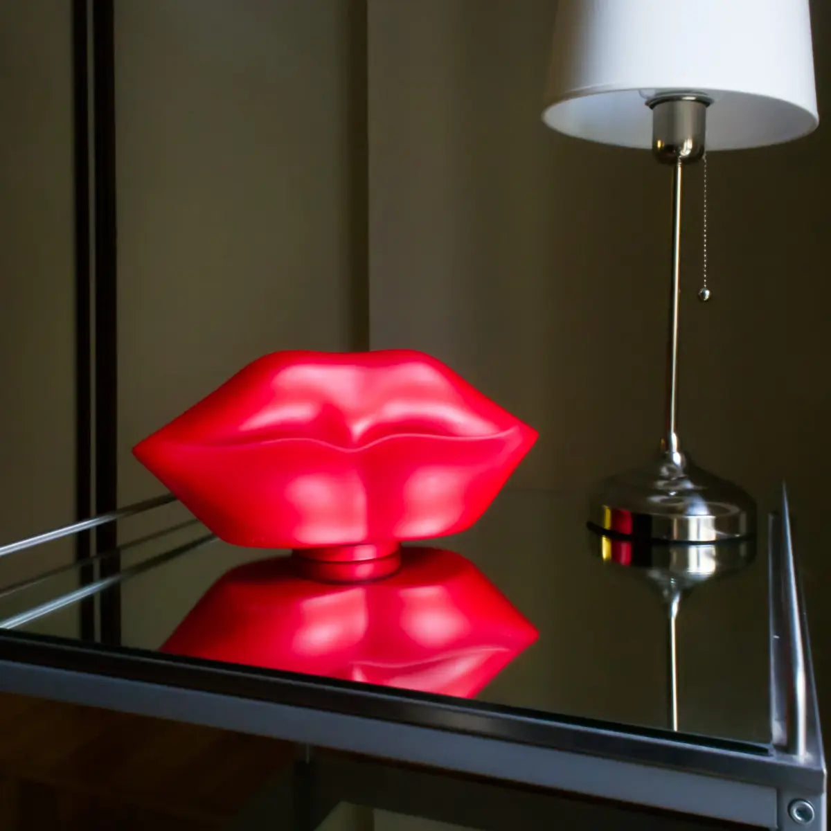 Lampada da tavolo rossa a forma di labbra. Appoggiata su un tavolino specchiato accanto ad una lampada con stelo argentato. Sfondo bianco