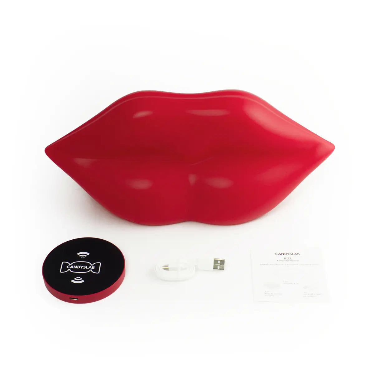 Lampada da tavolo rossa a forma di labbra. Appoggiati accanto alla lampada tutto ciò che contiene la confezione ovvero la base di ricarica wireless, il caricatore e il foglietto illustrativo. Sfondo bianco