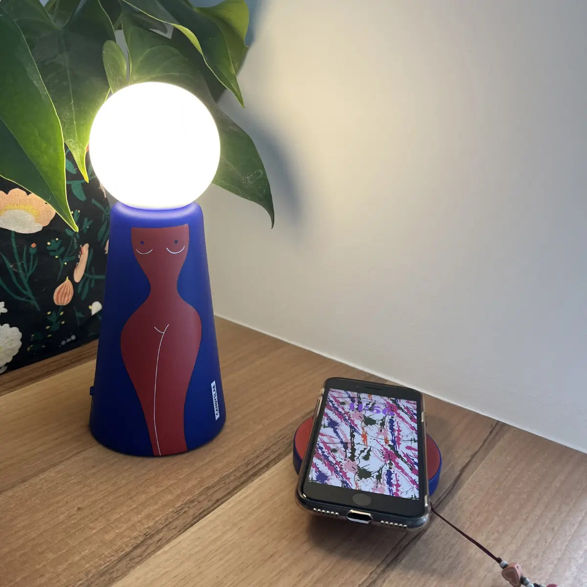 Lampada da tavolo Eva con base blu e figura femminile rossa posizionata su un tavolino di legno. Sulla base di ricarica è posizionato un telefono in carica.