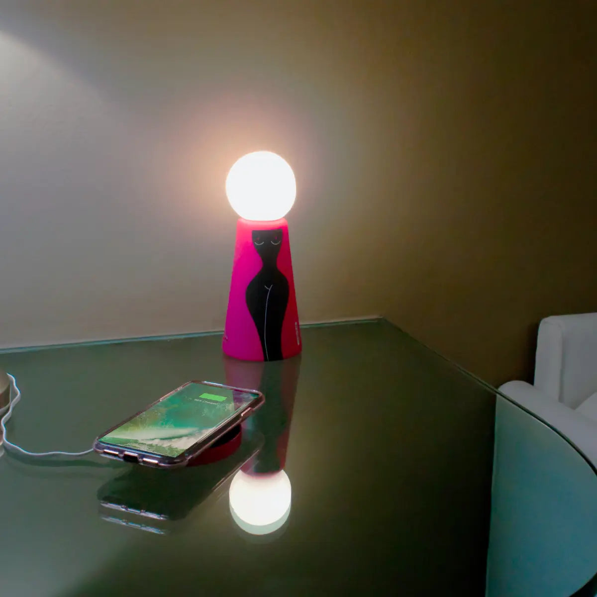 Lampada da tavolo Eva con base fucsia e figura femminile nera posizionata su un tavolino. Sulla base di ricarica è posizionato un telefono in carica.