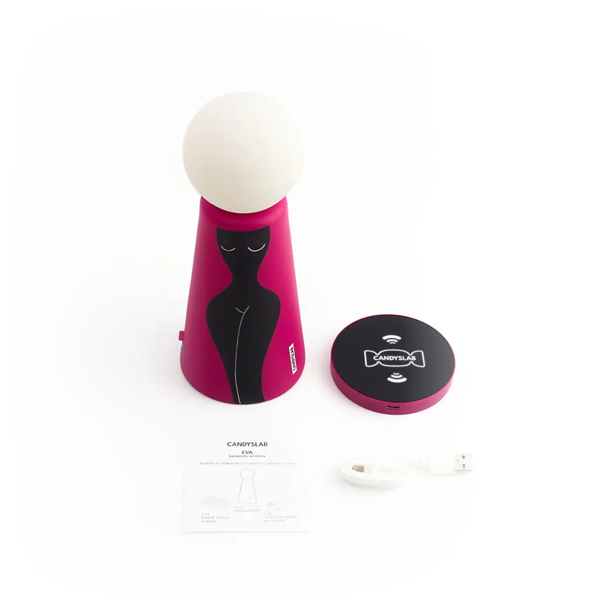 Lampada da tavolo a forma conica con sfera luminosa. Base fucsia com figura femminile nera. Posizionato accanto alla lampada la base di ricarica wireless, il cavo di ricarica e il libretto d'istruzione. Sfondo bianco 