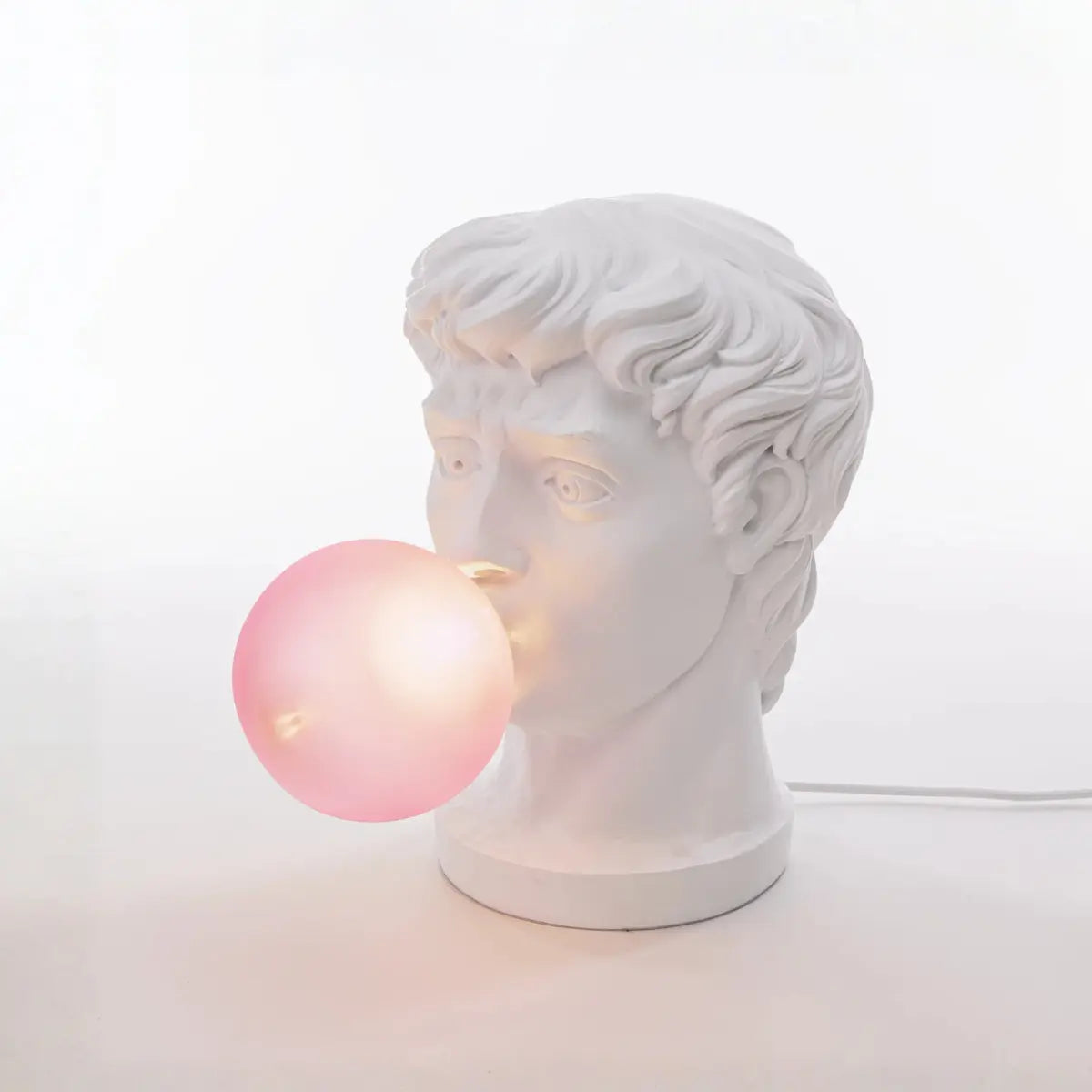 Lampada accesa di resina bianca che rappresenta il viso di un giovane ragazzo che fa una bolla di chewing-gum fucsia che diventa la lampadina. Sfondo bianco 
