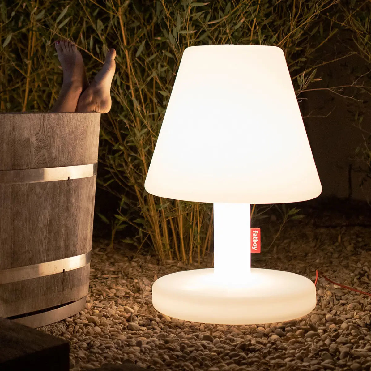 Lampada Edison The Grand di Fatboy bianca con dettaglio rosso posizionata all'esterno accanto ad una vasca in legno.