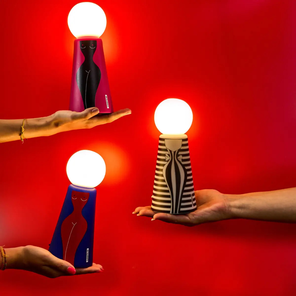 Tre mani con sopra appoggiate le tre versioni della lampada da tavola Eva. Le lampade sono accese e lo sfondo è rosso 