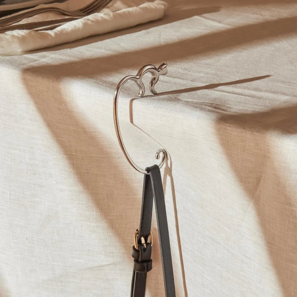 Minou, il gancio portaborse di Alessi a forma di gatto posizionato su un tavolo con tovaglia beige. Sul gancio è appesa una borsa nera.