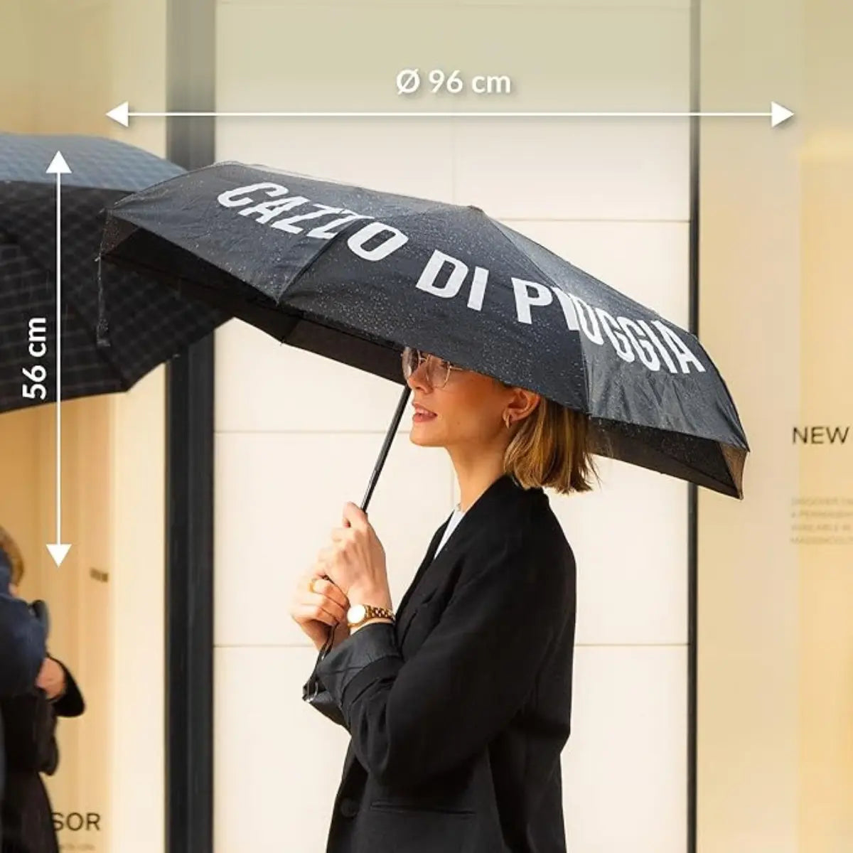 Donna vestita di nero che tiene in mano l'ombrello nero cazzo di pioggia. In bianco sono segnate le misure dell'ombrello da aperto. 