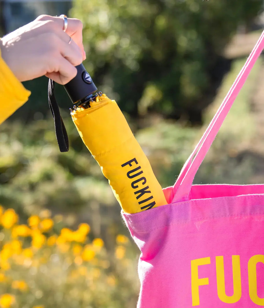 Mano che mette l'ombrello fucking rain giallo chiuso dentro una borsa rosa. Sfondo giardino