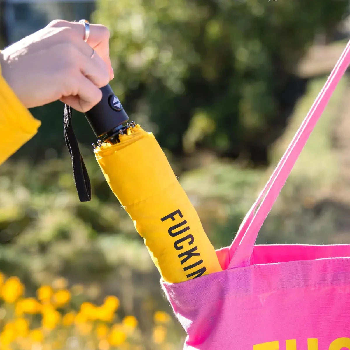 Mano che mette l'ombrello fucking rain giallo chiuso dentro una borsa rosa. Sfondo giardino