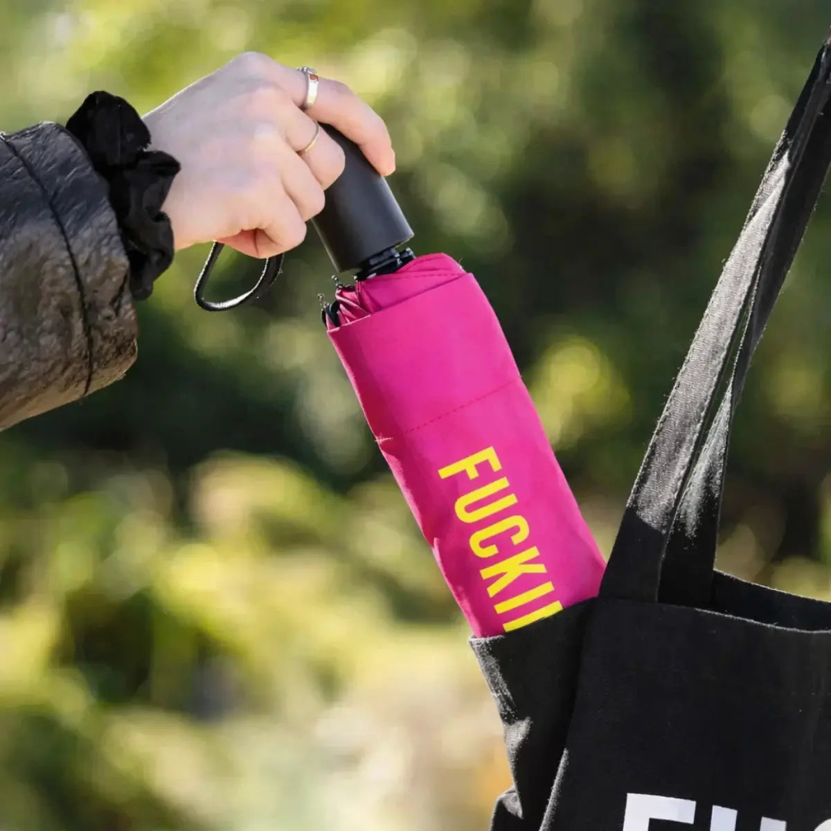 Mano che mette l'ombrello fucking rain fucsia chiuso dentro una borsa nera. Sfondo giardino
