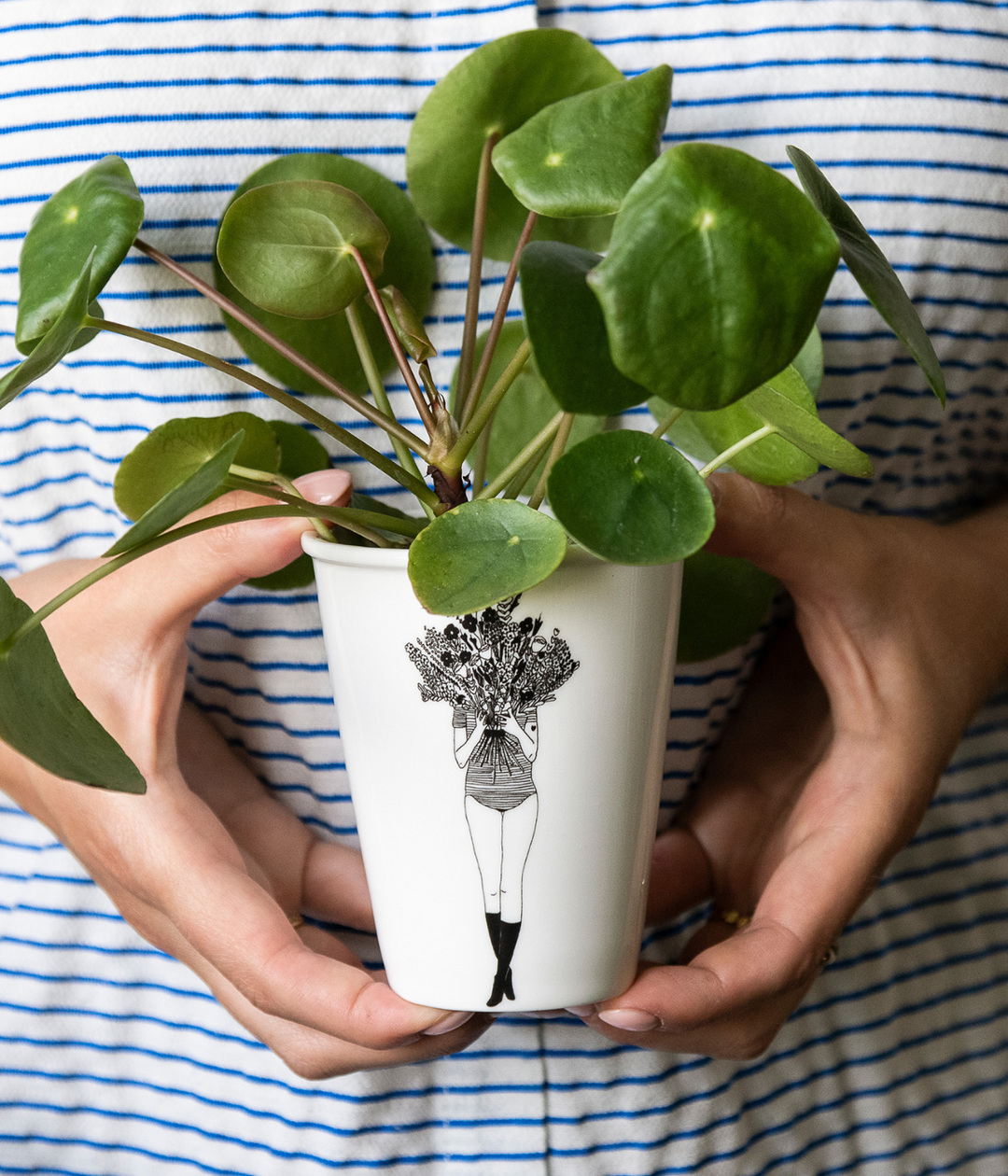 Mani che tengono una tazza bianca disegno nero e pianta verde all'interno. Maglia righe bianco blu.