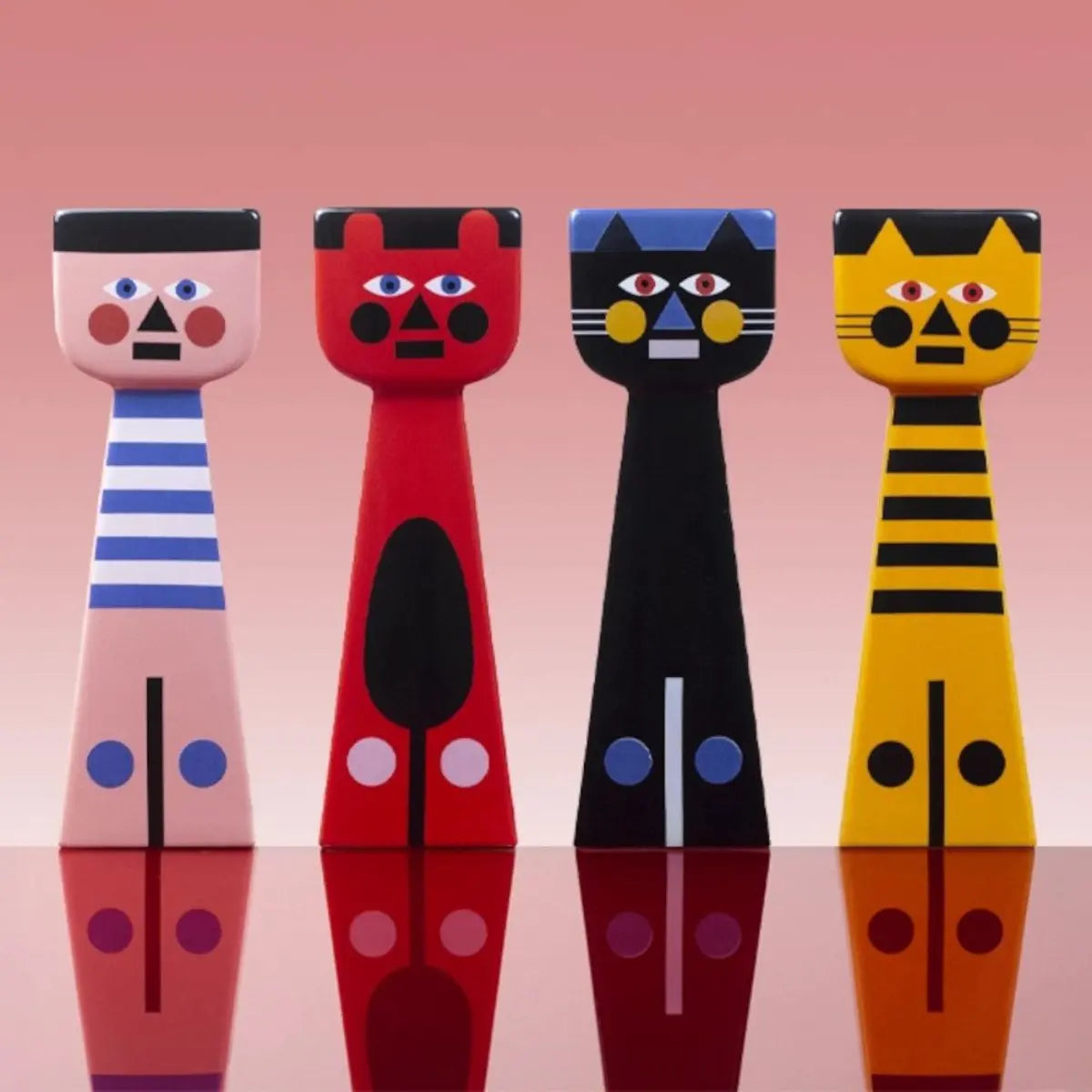 Quattro portacandele in ceramica di Qeeboo. Da sinistra: portacandele rosa con dettagli blu e bianchi, portacandele rosso con dettagli neri, portacandele nero con dettagli blu, gialli e bianchi, portacandele arancione con dettagli neri. Sfondo rosa sfumato