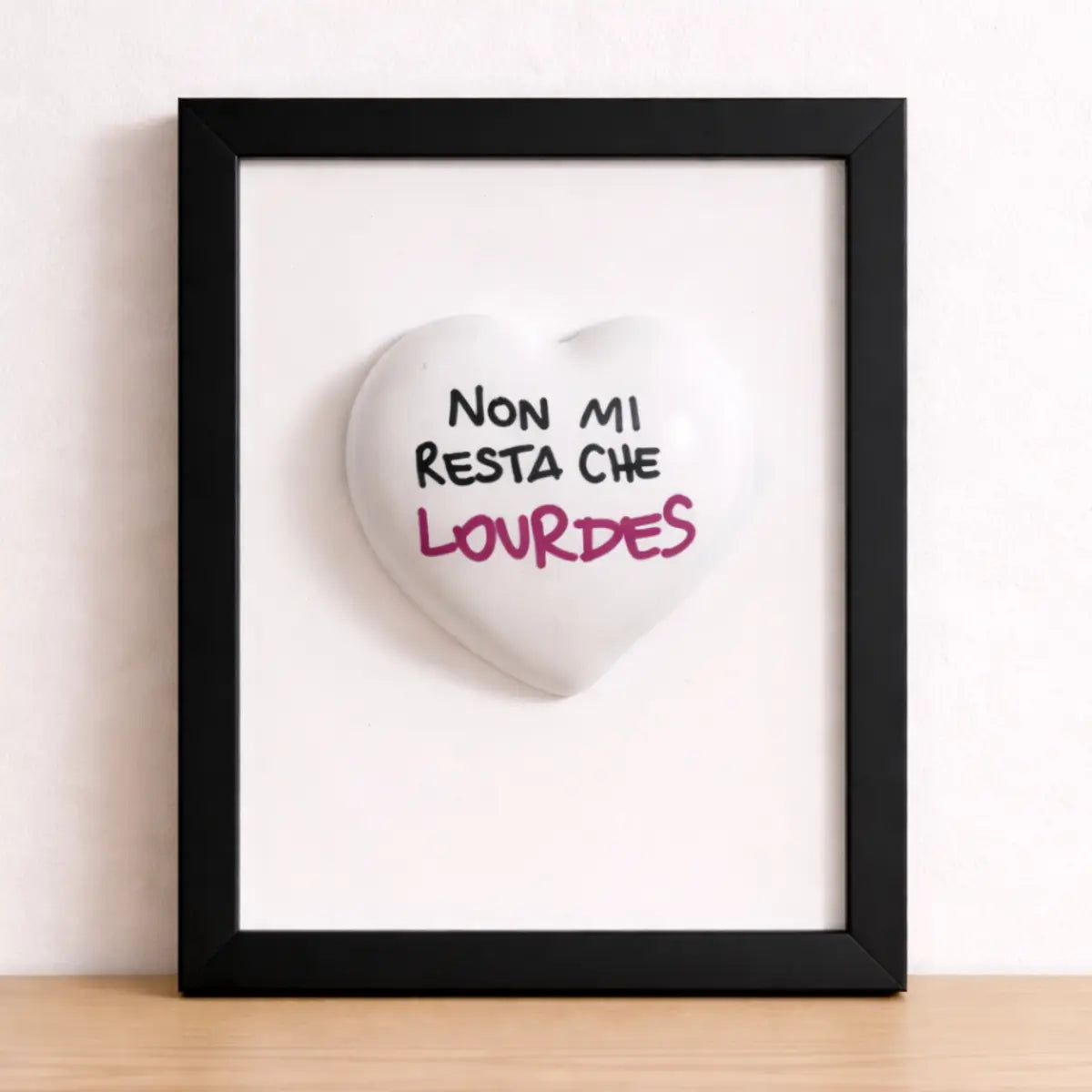 Quadretto con cornice nera, sfondo bianco e cuore bianco opaco con scritta "Non mi resta che Lourdes". Quadretto appoggiato su una mensola di legno