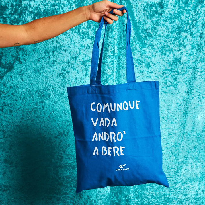 Una mano che tiene la shopper blu di Linea Daria con la scritta "Comunque vada andrò a bere"