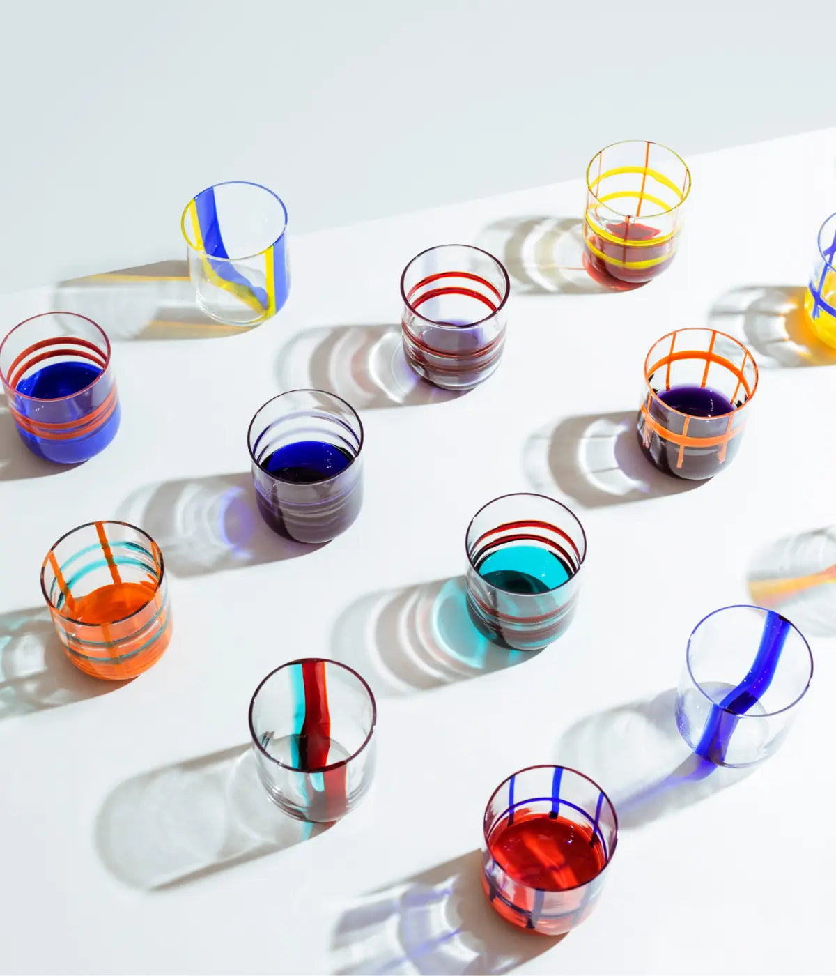 Bicchieri in vetro trasparente con diversi colori, righe e fondali. Appoggiati su tavolo bianco.
