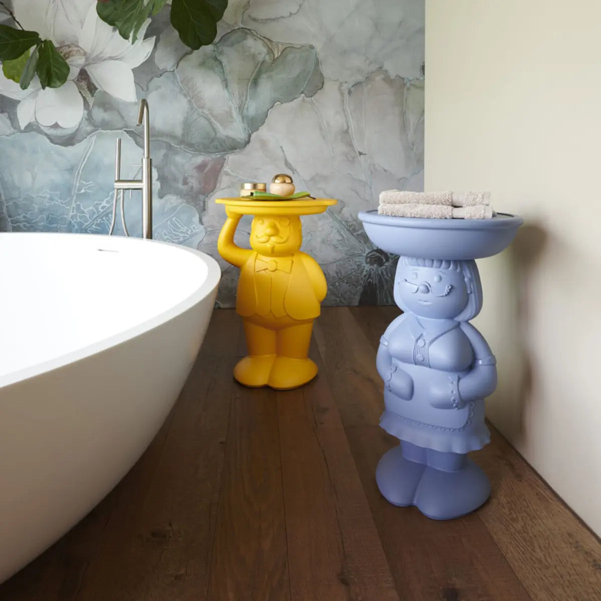 Un tavolino Ambrogio giallo e un tavolino Amanda celeste posizionati in un bagno accanto alla vasca da bagno. Pavimento parquet e carta a parati nel muro.