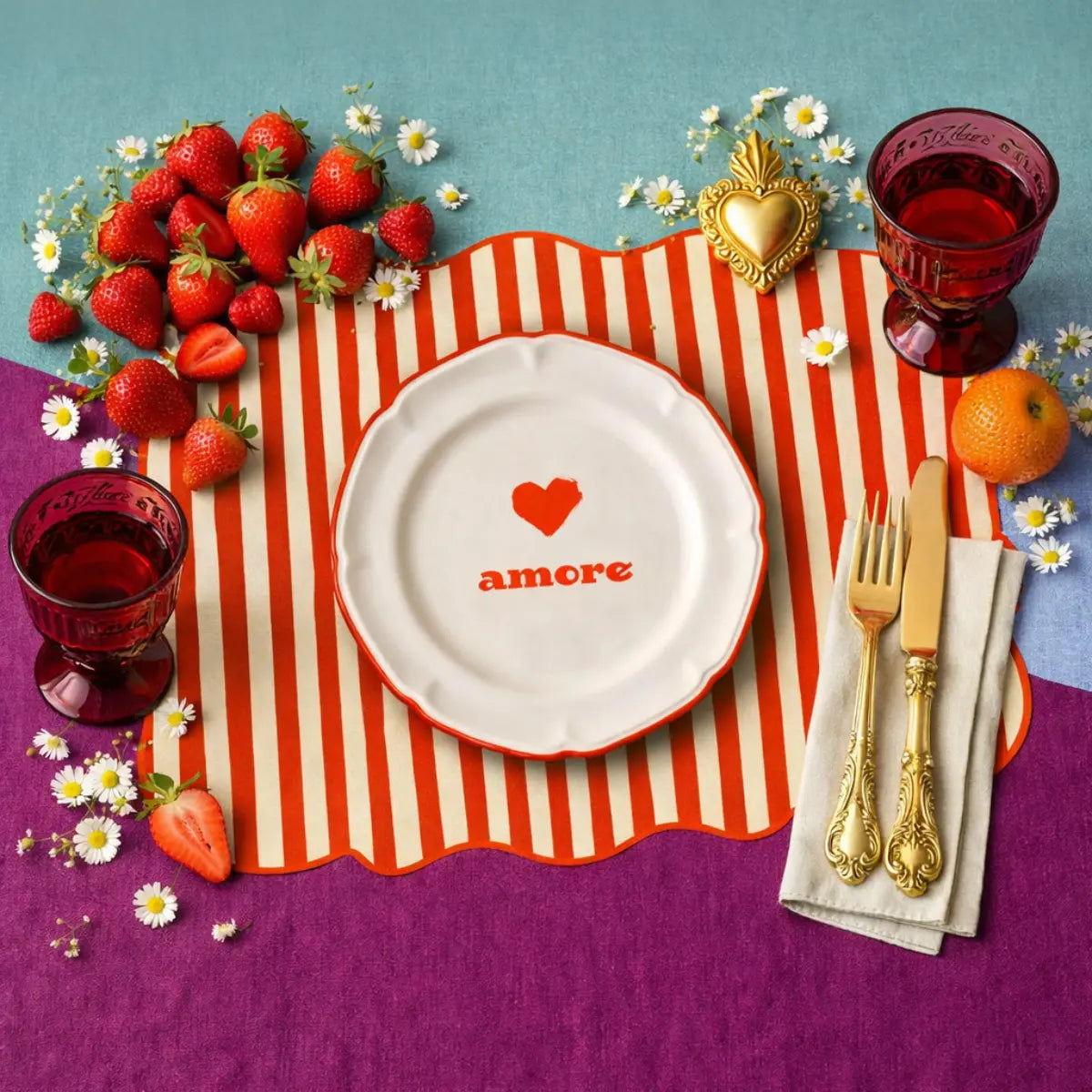 Tovaglietta dalla forma irregolare a righe bianche e rosse con al centro un disegno di un piatto bianco con la scritta "amore" e un cuore rosso disegnato.
La tovaglietta è appoggiata su un tavolo con delle fragole, dei fiori, dei bicchieri rossi e delle posate dorate appoggiate su un tovagliolo bianco. Lo sfondo è azzurro e viola