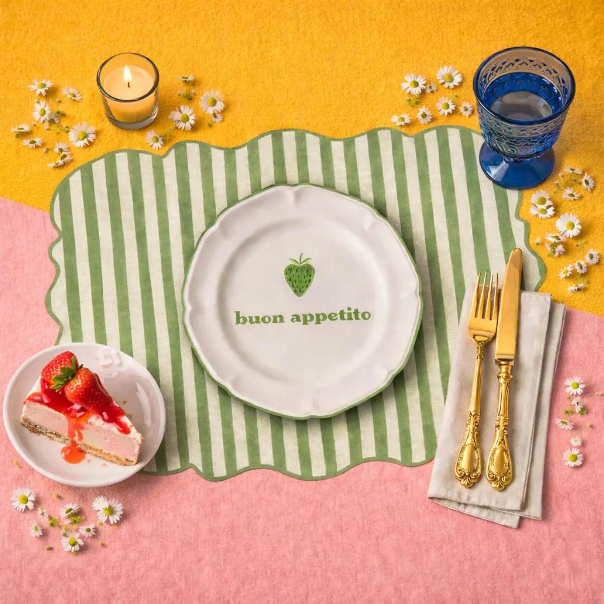 Tovaglietta dalla forma irregolare a righe bianche e verdi chiaro con al centro un disegno di un piatto bianco con la scritta "buon appetito" e una fragola verde disegnata. La tovaglietta è appoggiata su un tavolo rosa e giallo con appoggiato anche un piattino con una torta alle fragole, dei fiori, un bicchiere di vetro blu, una candeline bianca e delle posate dorate appoggiate su tovagliolo bianco