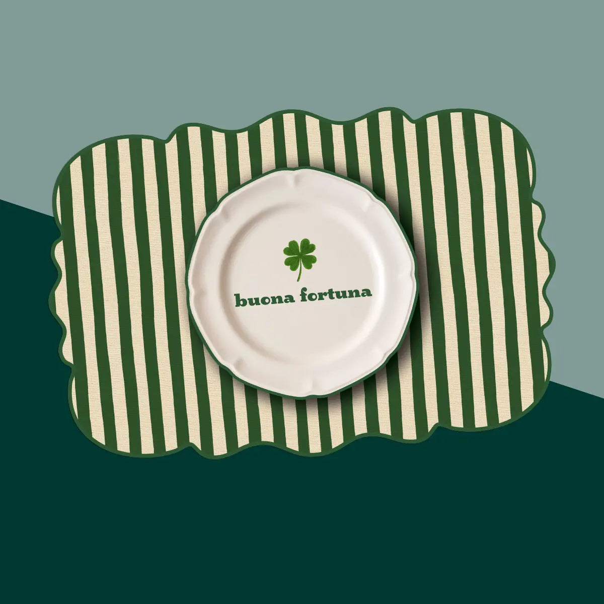 Tovaglietta dalla forma irregolare a righe bianche e verdi con al centro un disegno di un piatto bianco con la scritta "Buona fortuna" e un quadrifoglio verde. Sfondo bicolore, diviso secondo una linea diagonale. Lo sfondo è verde scuro e un verde acqua più chiaro.