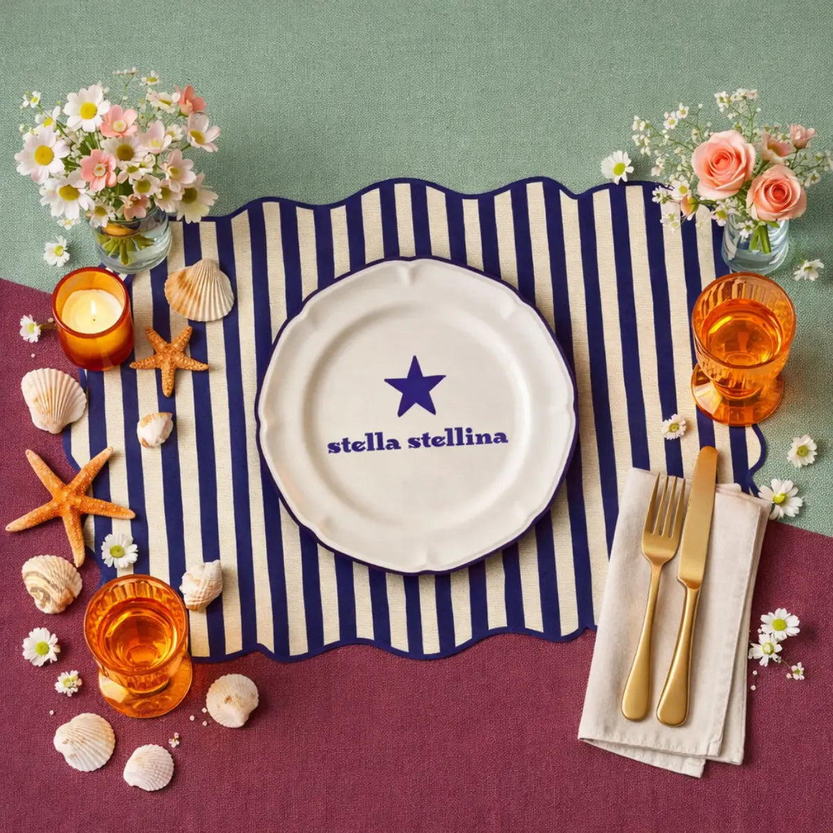 Tovaglietta dalla forma irregolare a righe bianche e blu con al centro un disegno di un piatto bianco con la scritta "stella stellina" e una stella blu disegnata. La tovaglietta è apoggiata su una tavola con sfondo rosa/viola e azzurro, diviso in diagonale. Oltre alla tovaglietta sul tavolo sono appoggiate delle conchiglie e stelle marine, dei bicchieri di vetro arancione, una candeline, dei fiori e delle posate dorate appoggiate su un tovagliolo bianco
