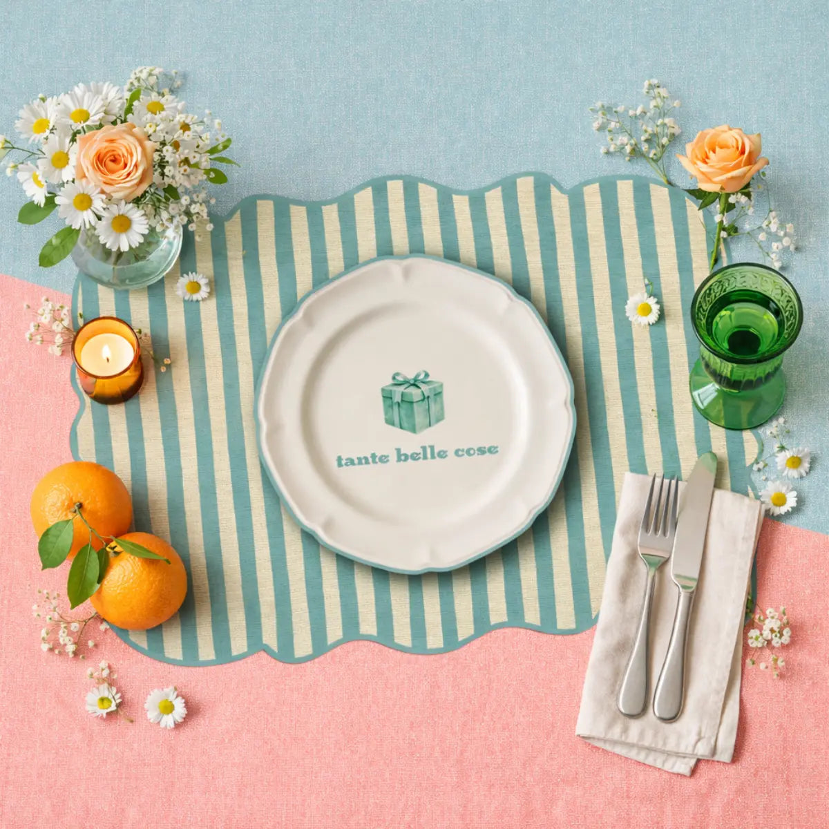 Tovaglietta dalla forma irregolare a righe bianche e azzurre con al centro un disegno di un piatto bianco con la scritta "tante cose belle" e un pacco regalo azzurro disegnato. La tovaglietta è appoggiata su una tavola. Oltre alla tovaglietta sul tavolo sono presenti dei mandarini, una piccola candela, dei vasetti di fiori e dei fiori sparsi, un bicchiere di vetro verde e delle posate argentate appoggiate su un tovagliolo bianco. Lo sfondo è diviso in due blocchi colorati, uno rosa chiaro e l'altro azzurro.