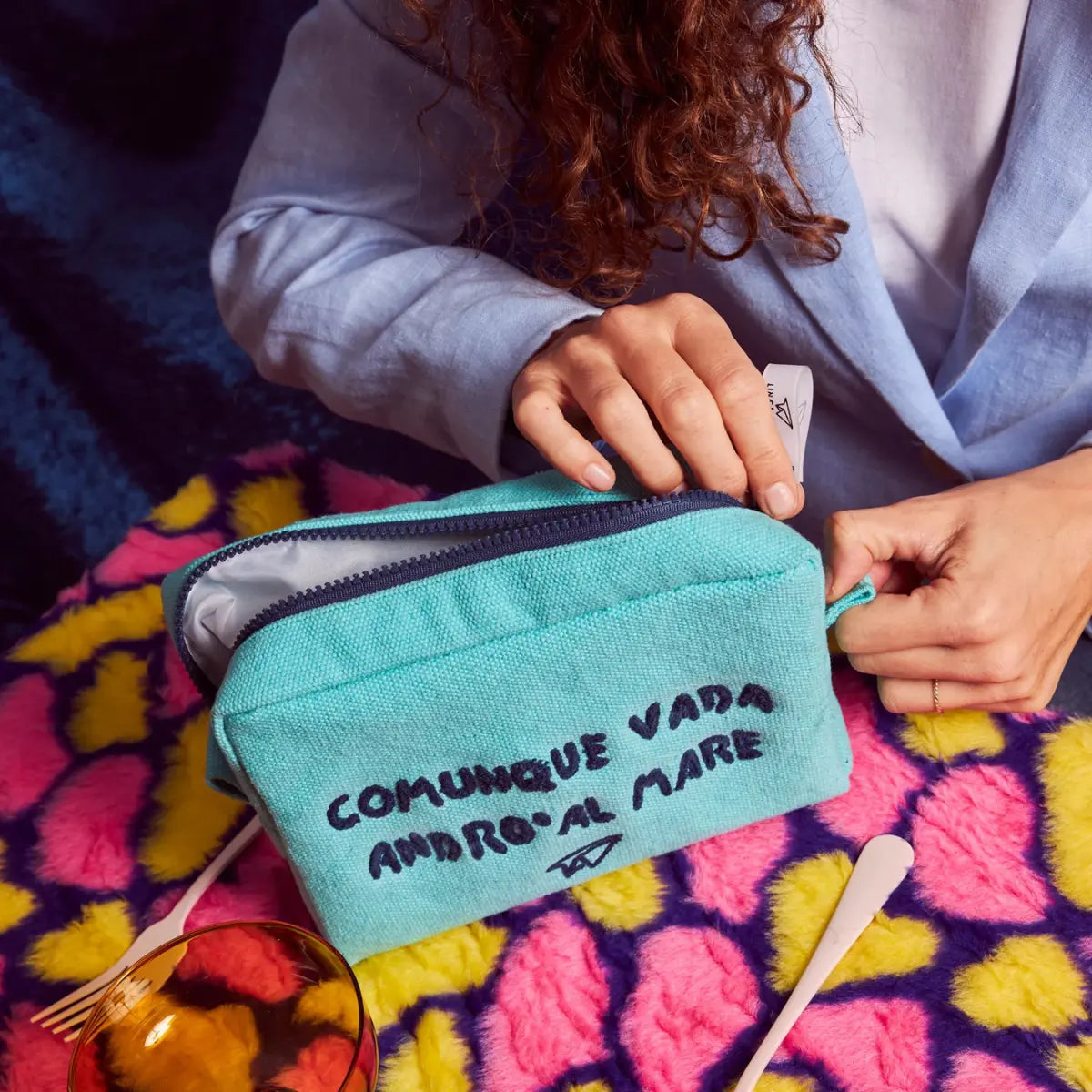 Mano che apre la zip blu della trousse azzurra con la scritta blu "Comunque vada andrò al mare"